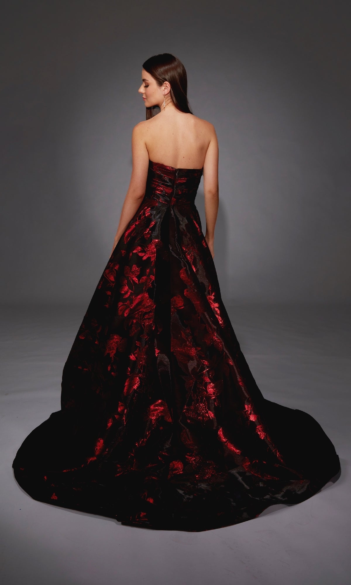 A woman stands facing away, wearing the Alyce 27862 long formal dress—a strapless, floor-length black and red floral jacquard gown with a sweeping train—against a plain dark background.